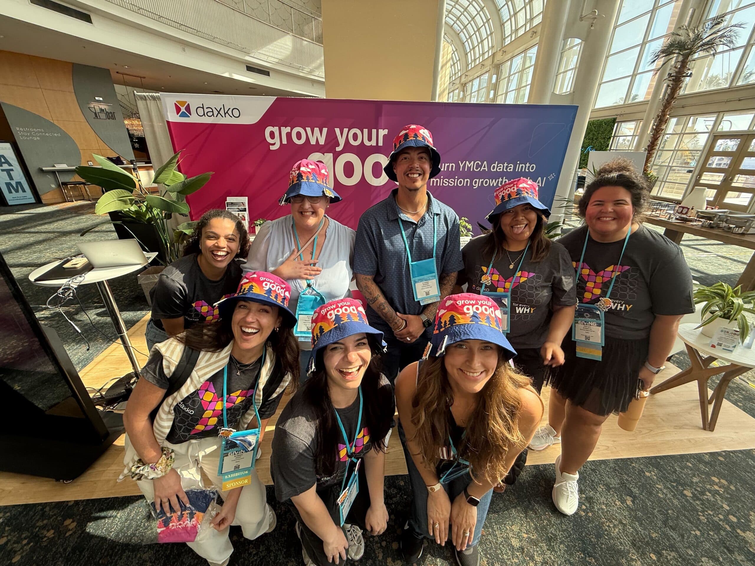 Eight smiling people pose at the Daxko booth at NAYDO, wearing “grow your good” hats.