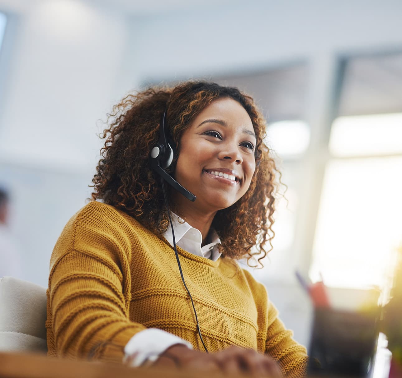 A smiling customer support representative wearing a headset works at a computer in a bright office.